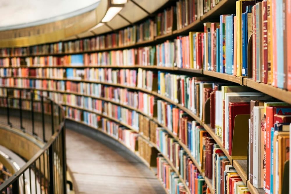 Tumpukan buku-buku tebal dan kacamata riset di perpustakaan universitas